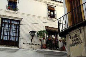 White building with wrought-iron balconies and potted plants; signs read 'Doña María Coronel' and 'Busostavera' on the façade.